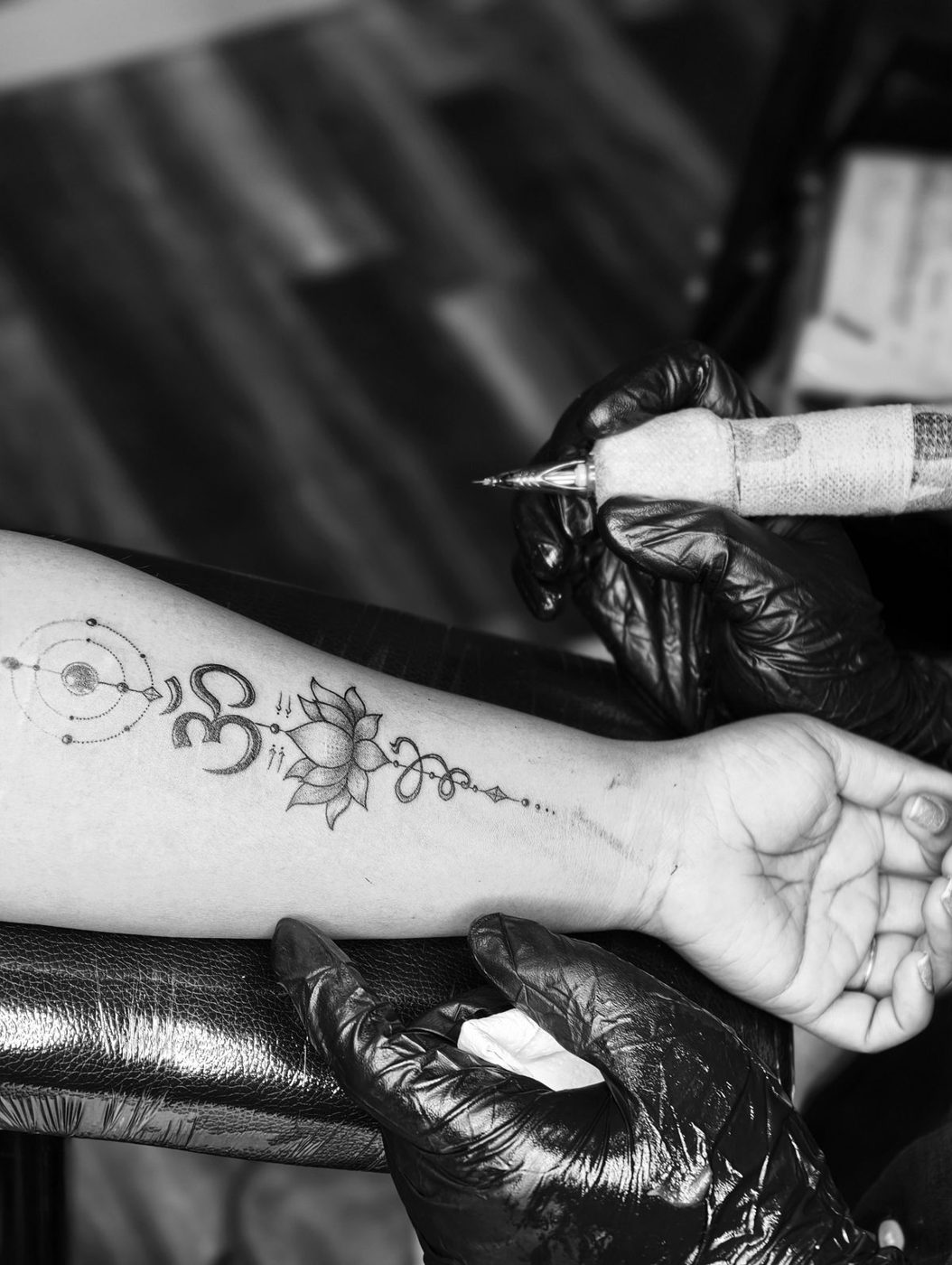 Black-and-white photo of a gloved tattoo artist working on a fine-line symbol design along a forearm