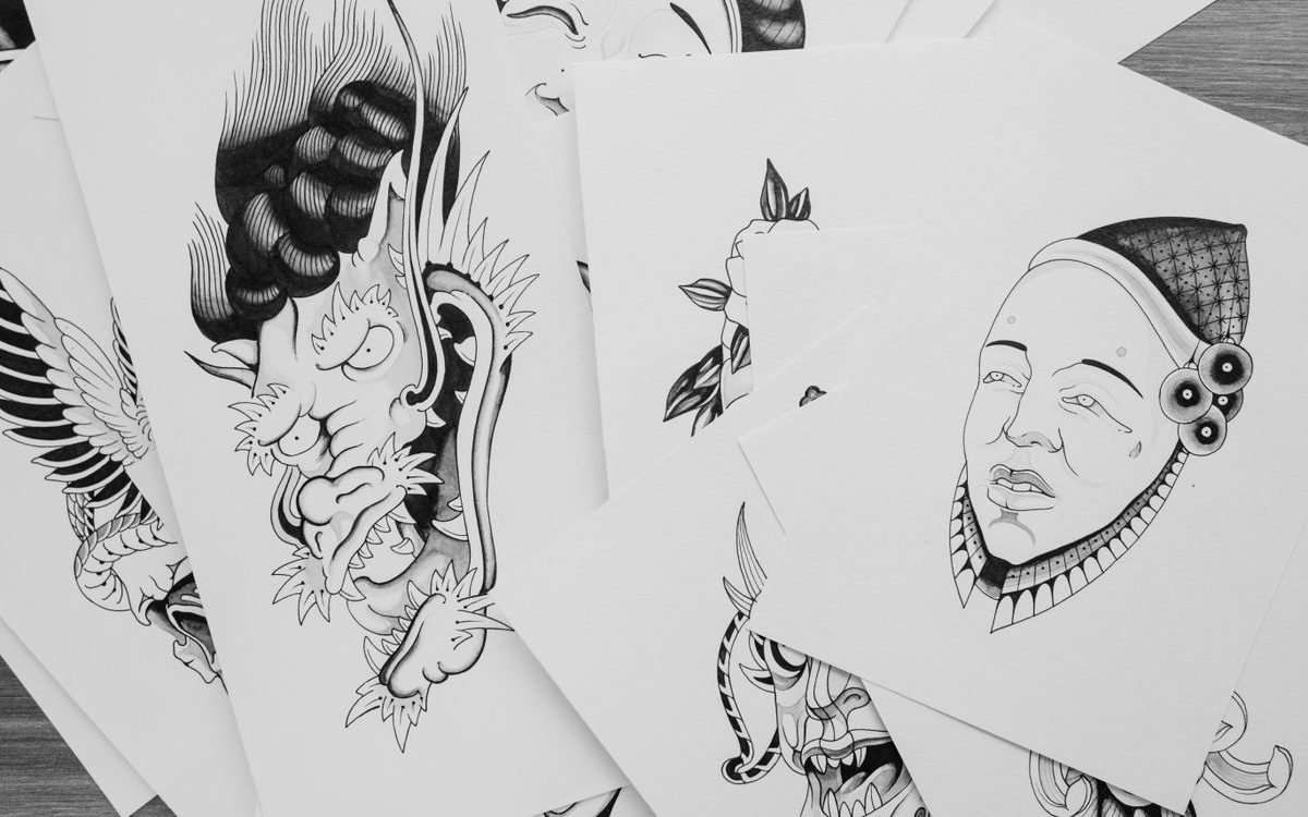 Sheets of black ink tattoo flash drawings, including a horned demon and a masked face, spread on a table