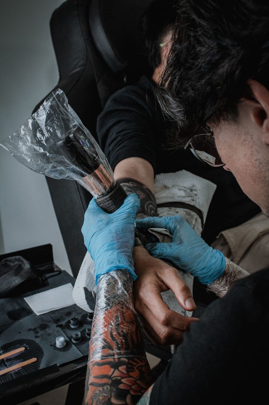 Tattoo artist in blue gloves holding clear wrap over a client's tattooed forearm