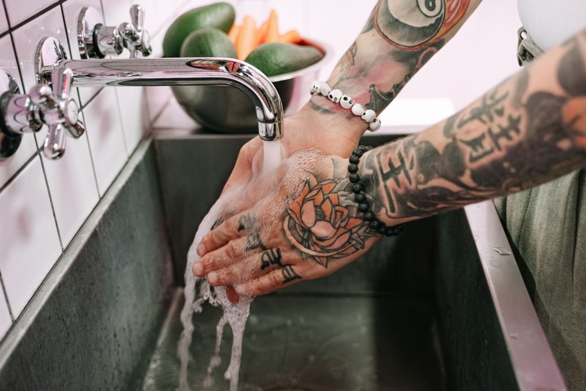 Tattooed hands and forearms being washed under a running tap