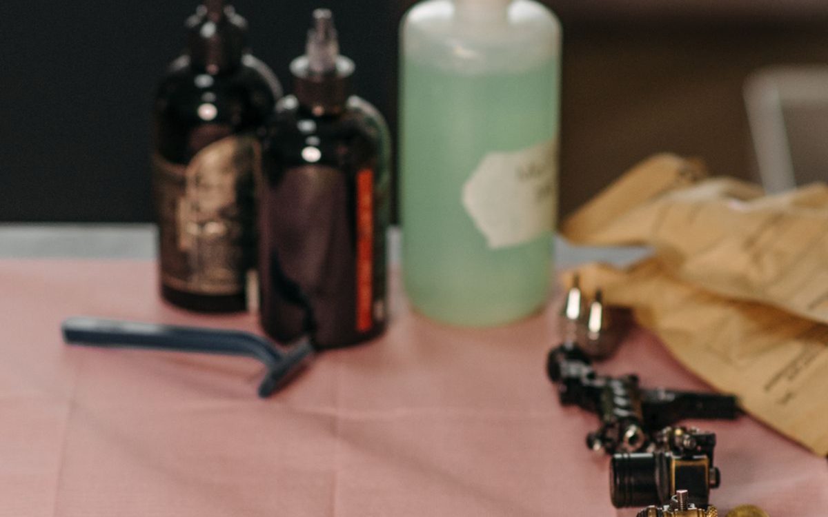 Tattoo supplies laid out on a pink barrier sheet: ink bottles, a wash bottle, sealed needle pouches and machines