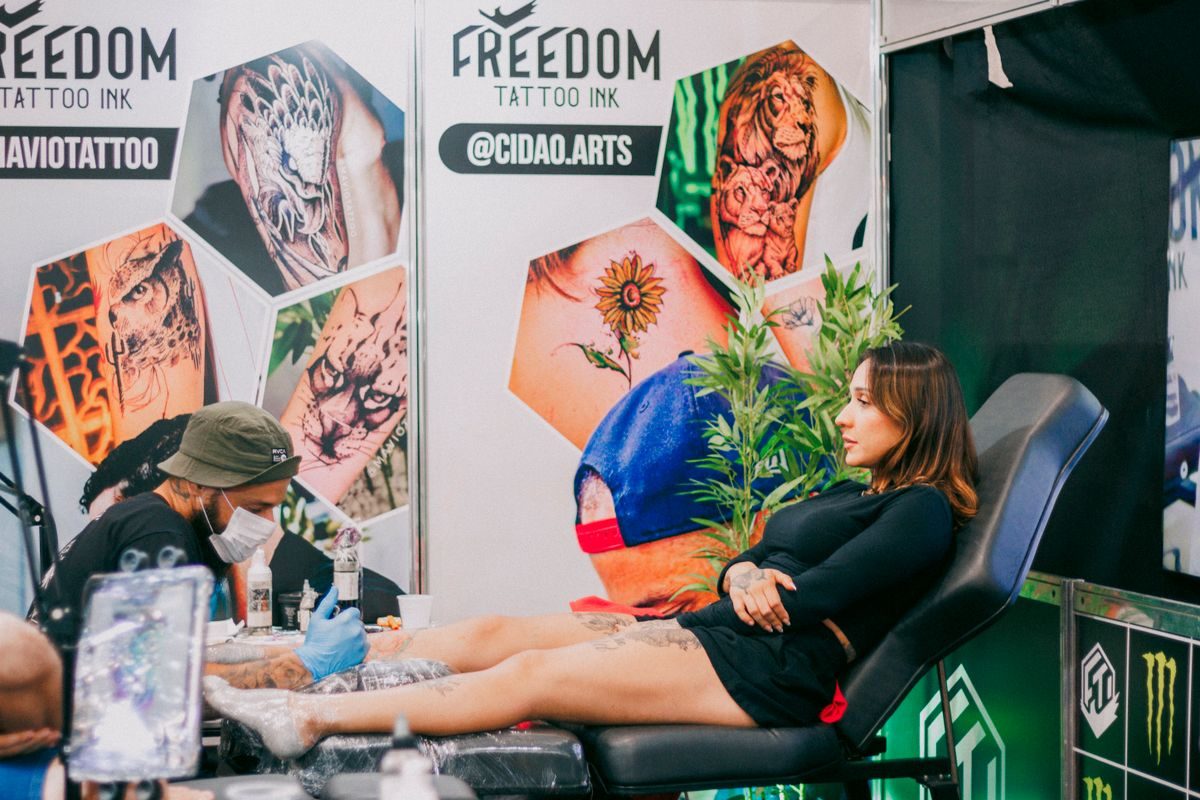 Tattoo artist working on a client's leg inside a convention booth hung with banners and flash