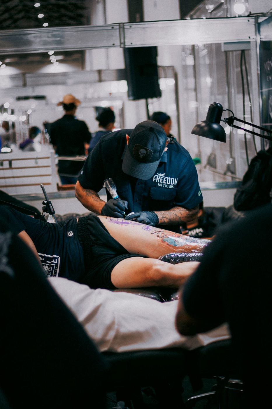 Artist in a cap tattooing a client lying on a table in a busy convention hall