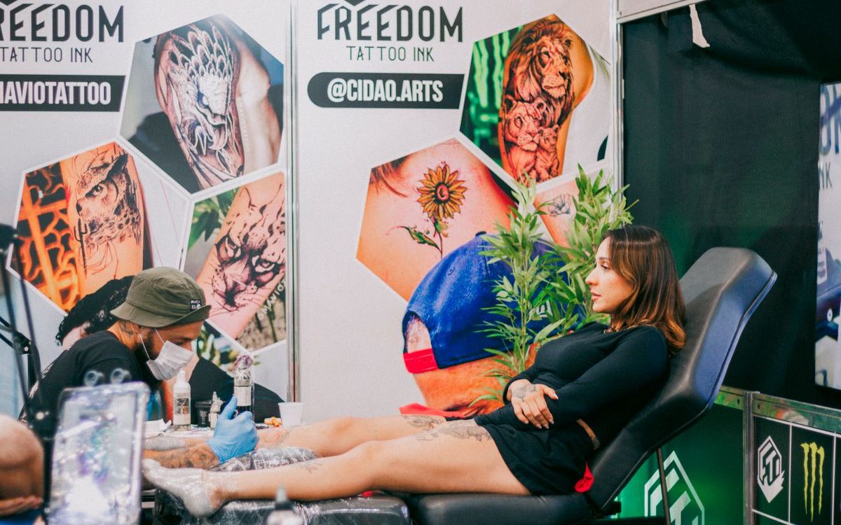 Tattoo artist working on a client's leg inside a convention booth hung with banners and flash