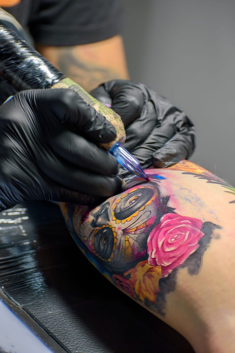 Artist in black gloves tattooing a colorful design with a pink rose and bold outlines on a leg