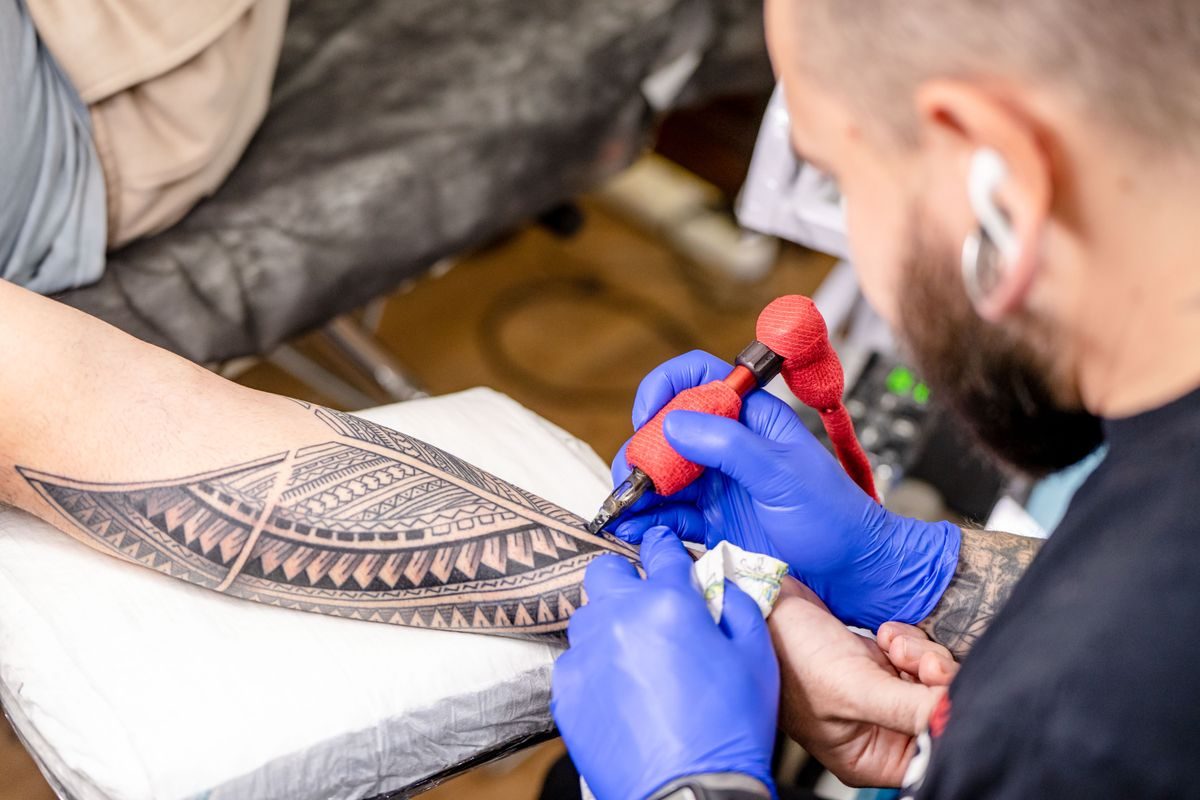 Artist in blue gloves tattooing a bold black geometric pattern along a forearm